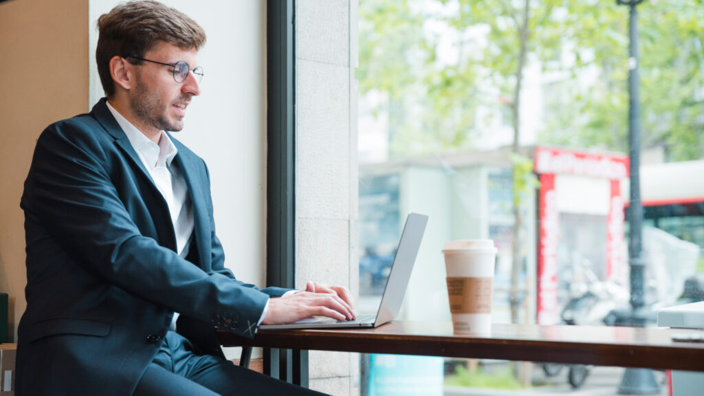 A man wearing a suit and glasses sits in the window of a cafe on his laptop, beginning the application for his Lithuania business visa