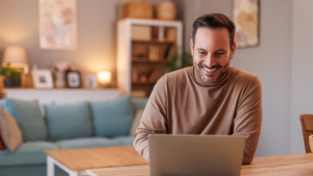 A young male wearing a beige jumper sits at his living room table reading an article on his laptop about the Wise business account
