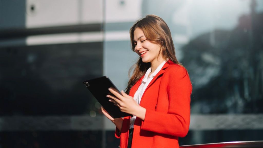 A young brunette female in a red suit  stands outside and smiles as she reads an article on her ipad about 'Is Wise Business safe?'