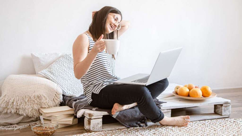 A young brunette female wearing glasses, jeans and a striped sleeveless top sits on a upcycled crate next to cushions and blankets as she holds a mug of coffee and reads an article on a laptop on her knee about how to pay international employees