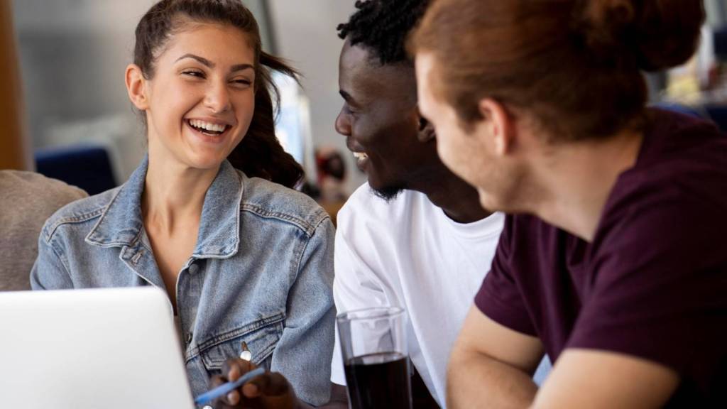 A young brunette female in a denim jacket and two young men wearing T-shirts talk and laugh to each other as they discuss the process of how to pay international employees