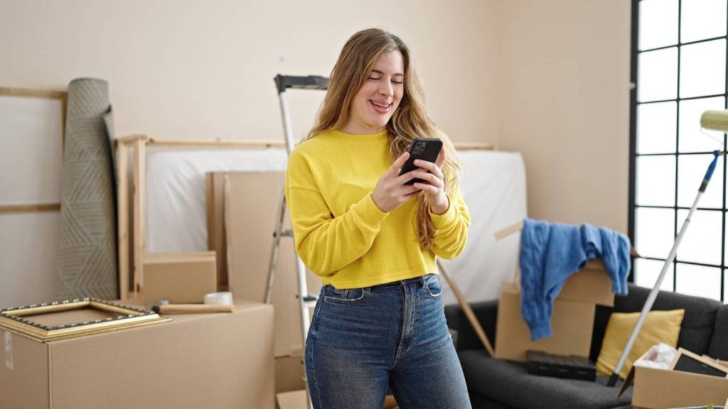A young woman with long hair stands in a room filled with boxes and decorating equipment. She wears a yellow top and blue jeans and smiles as she reads about the Revolut Business Account on her mobile phone