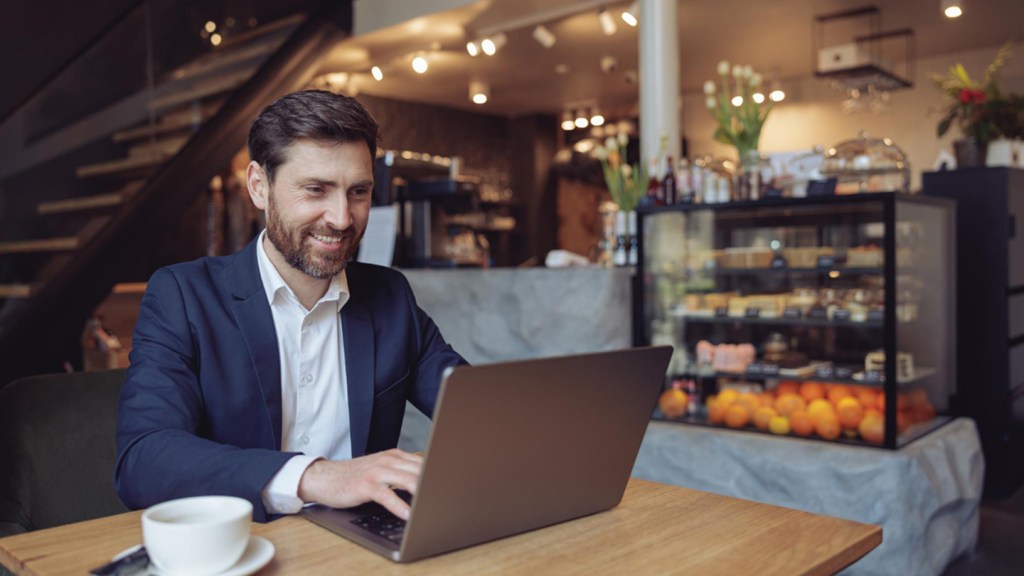 A man with a beard dressed in a smart suit and shirt sits at a table and reads an article on his laptop about digital payment platforms