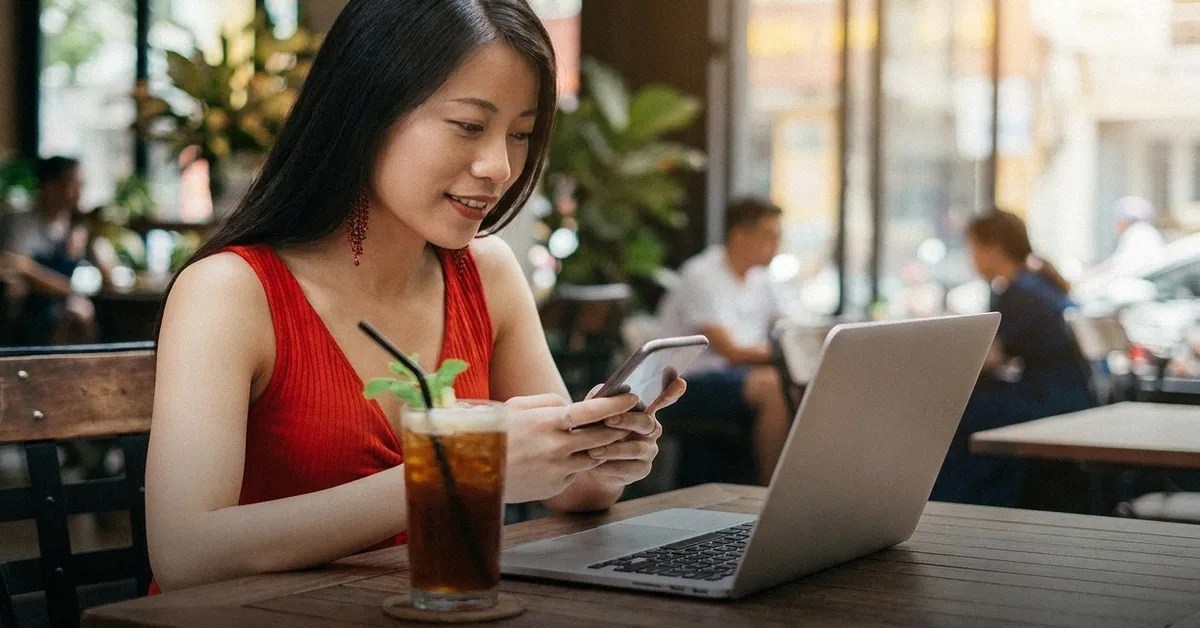 A young woman sits in a cafe with a drink and her laptop on the table while reading about buying a property in the Philippines on her mobile phone