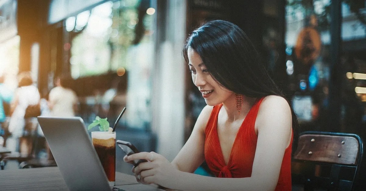 A young woman sits in a cafe with a drink and her laptop on the table while reading about buying a property in the Philippines on her mobile phone