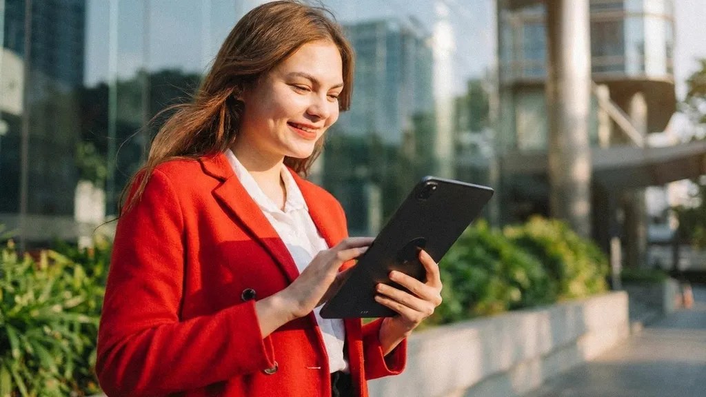 A young brunette female in a red suit  stands outside and smiles as she reads an article on her ipad about 'Is Wise Business safe?'