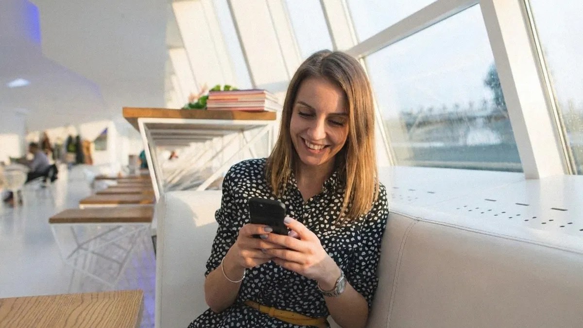 A young brunette female in a patterned shirt sits on the sofa and reads an article on her phone about how to pay international contractors