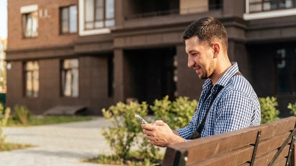 A young man in a blue check shirt looks ahead as he sits on a bench outside and reads about the Revolut Business Account on his mobile phone
