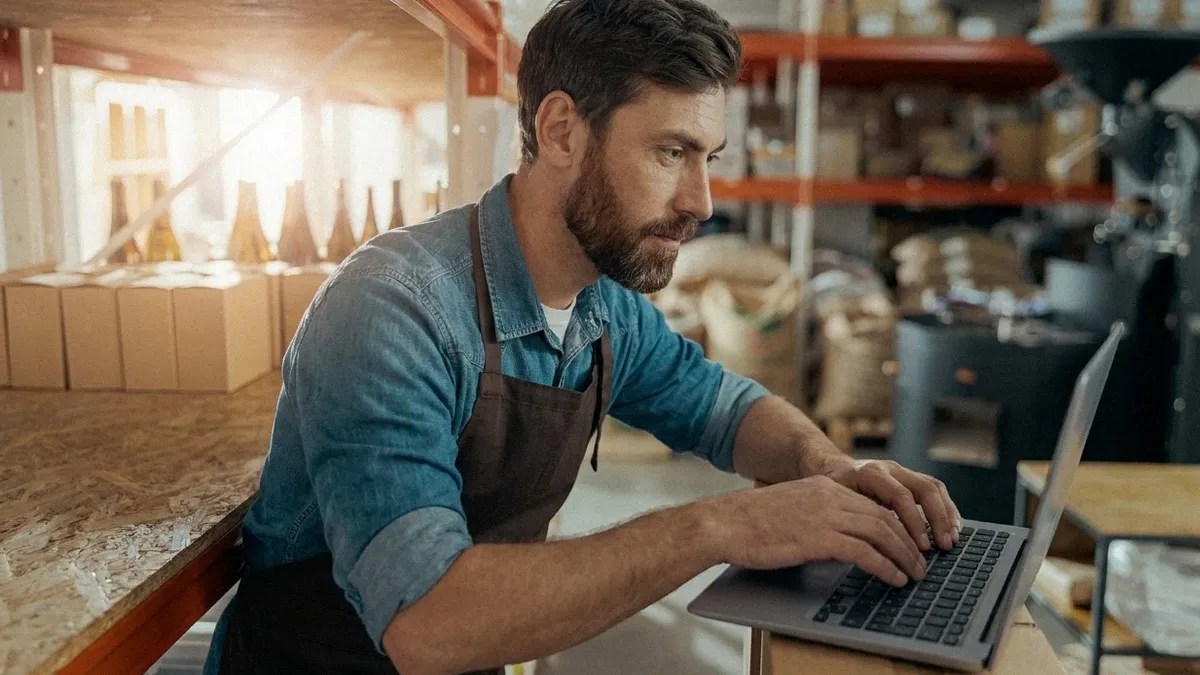 A young bearded man in a blue shirt and apron stands up in a cafe while typing on his laptop to simplify his supplier payments process