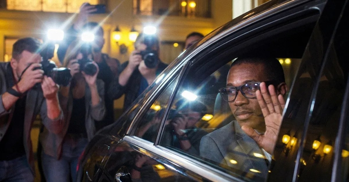 A man in a car waves to people outside as people take photographs