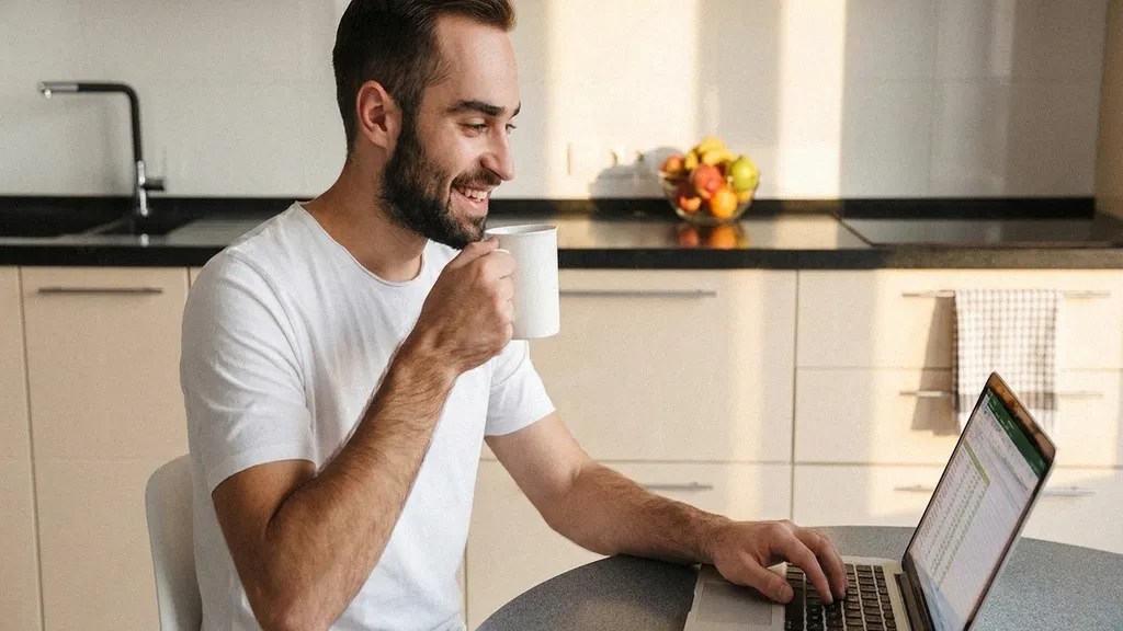 A man in a white T-shirt sits at a desk reading an article on his laptop about the benefits of TransferGo vs Wise Business